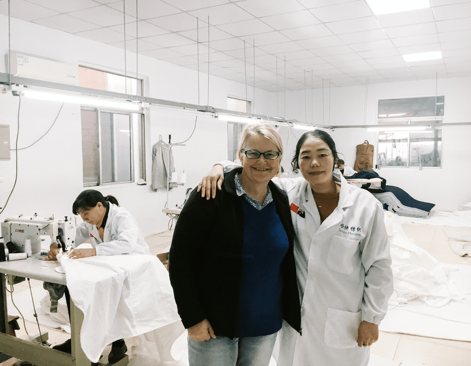Diane and Factory worker in the sewing room at the factory.  In the background a working at a sewing machine is working on a White Brolly Sheet.