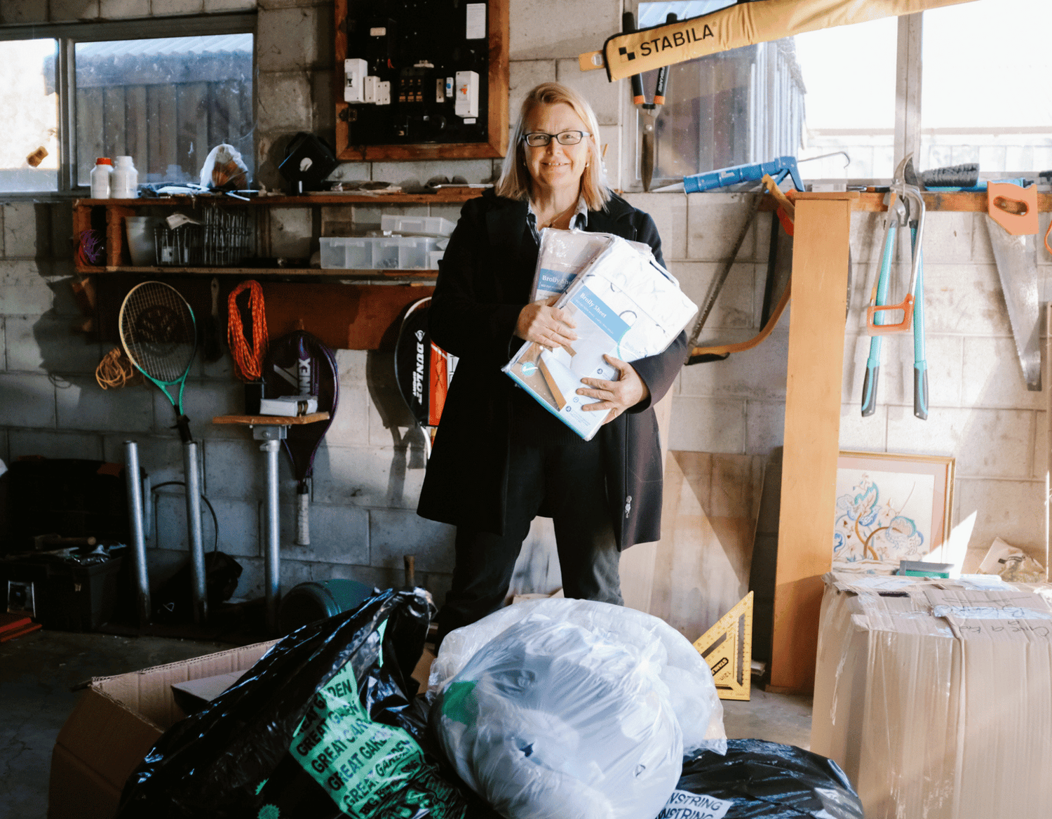 Early photo of Diane in garage sorting through delivery of various materials in black rubbish sacks, while holding 2 packaged Brolly Sheets.