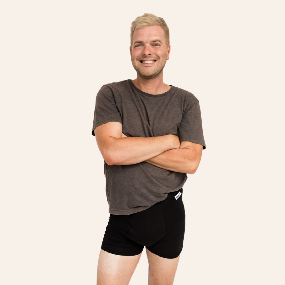 A smiling man with blonde hair stands with arms crossed, wearing a dark brown short-sleeve t-shirt and black ultra absorbent underwear against a solid off-white background.