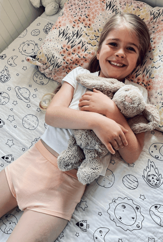 Happy young girl wearing our snazzi pants organic night trainers on her bed cuddling her teddy