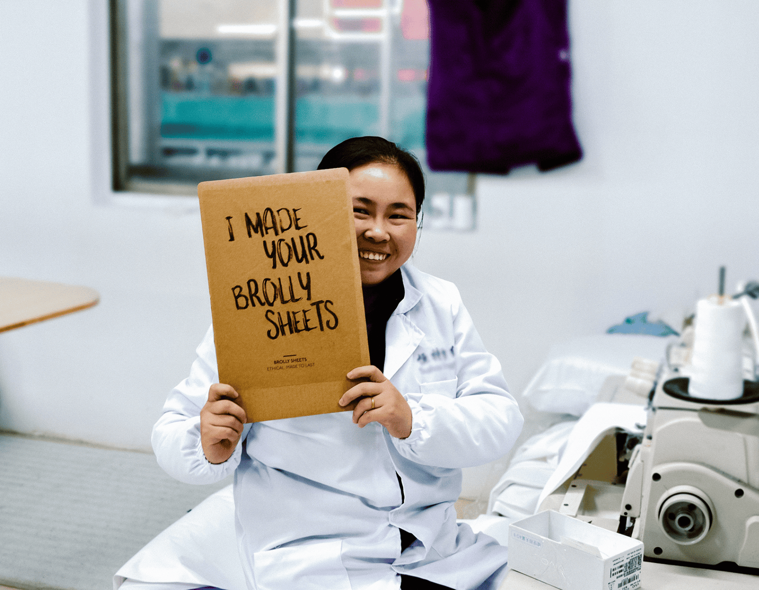 Factory worker at sewing machine holding up a sign with the words "I made your Brolly Sheets"
