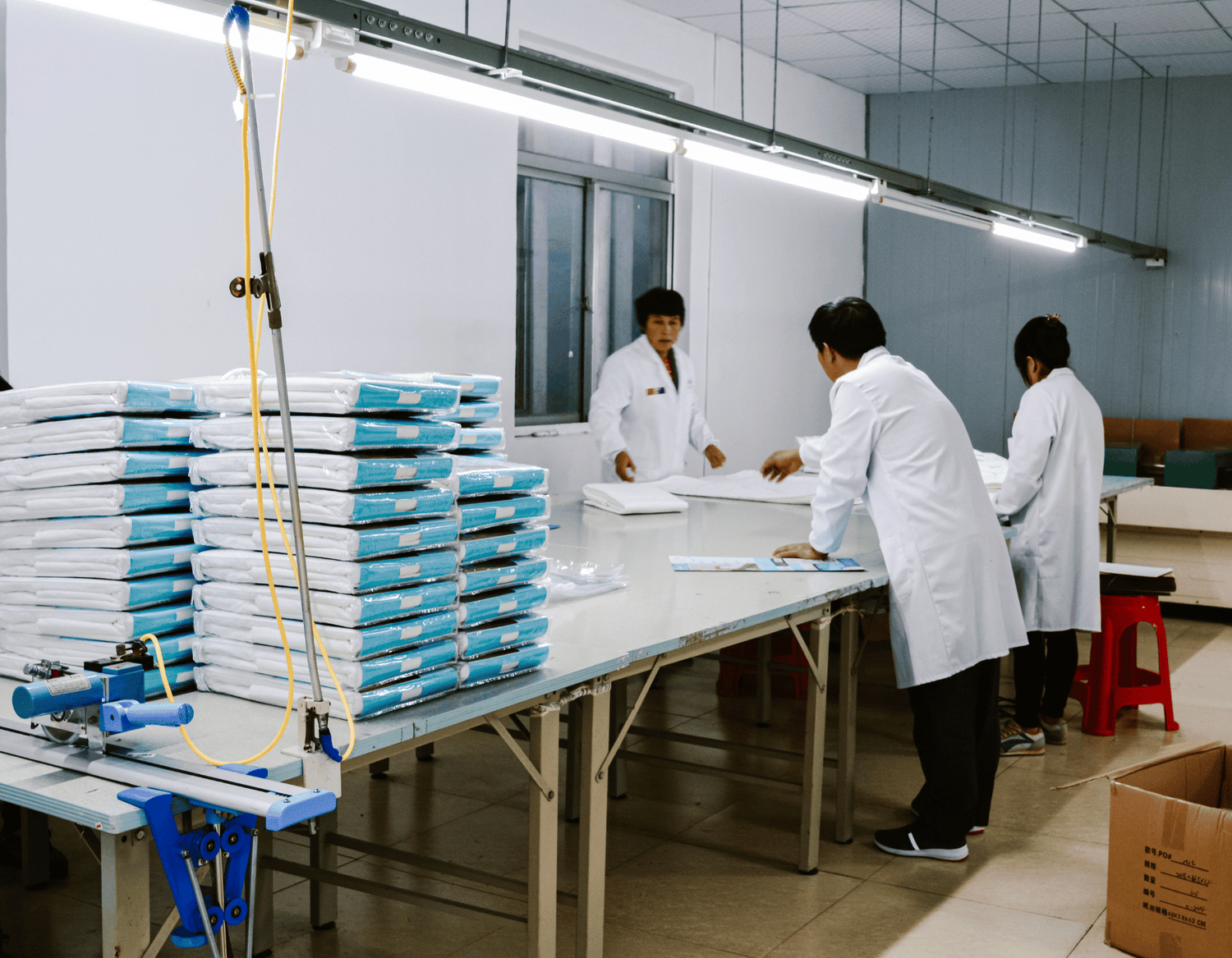 Workers in factory folding Brolly Sheets on long white table.  In the foreground there are 5 stacks of Brolly Sheets waiting to be put in to cartons.