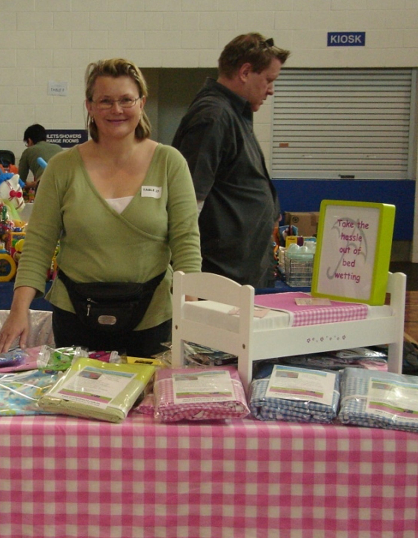 Diane standing in front of table with pink gingham table cloth.  on the table are several brolly sheets a and a scale model bed with scale model pink brolly sheets.  the sign says take the hassle out of bed wetting.