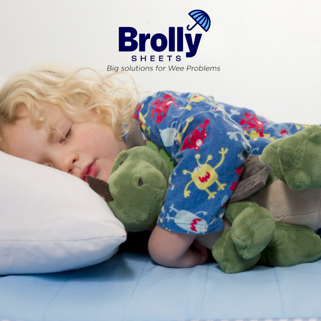 Child in colorful pajama pants with cartoon designs lying on a bed with green dinosaur pillows, featuring Brolly Sheets