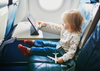 Toddler in airplane seat, holding a model aero plane while looking at ipad that is resting on on the folded table table from the seat in front her.