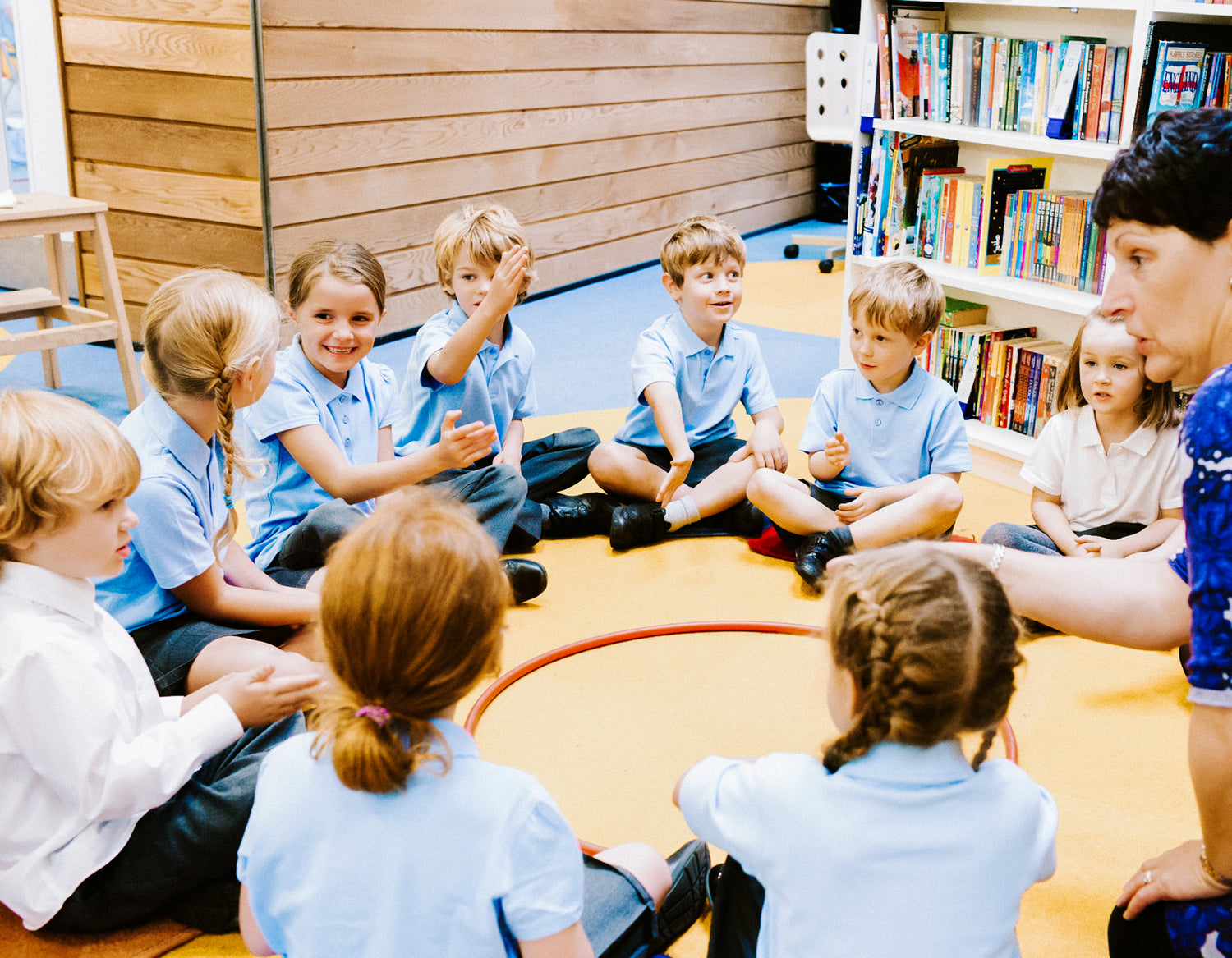 School children, sitting on a mat in a circle around a hula hoop.  3 shelves of library books can be seen in the background.